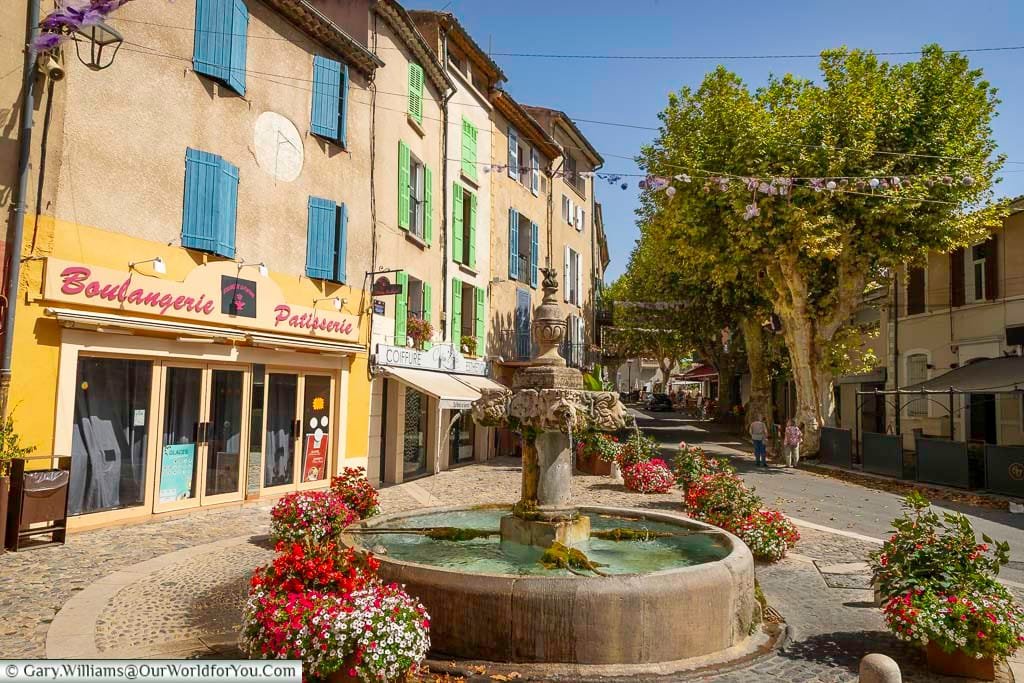Charming village square in Provence featuring a historic stone fountain surrounded by vibrant flower displays, with colorful shutters and a traditional boulangerie. This picturesque scene captures the authentic charm and daily life of southern France’s most beautiful villages.