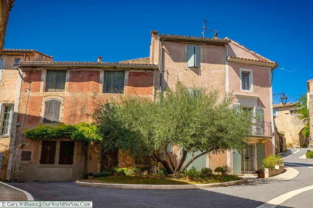 A peaceful village corner in Provence features pastel-hued houses with rustic shutters and a sunlit olive tree at the center of the quiet square. This charming Provençal scene captures the timeless beauty and relaxed atmosphere of southern France, perfect for travelers seeking authentic French countryside experiences.