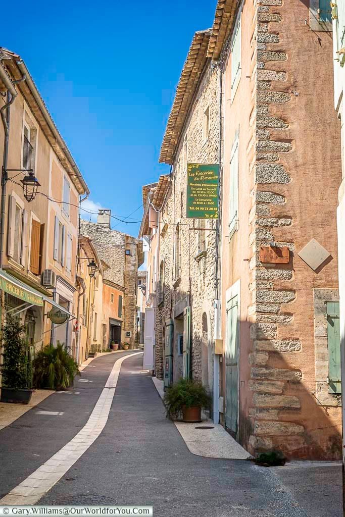 A peaceful, sunlit street winds through a charming Provençal village, lined with pastel-hued buildings, green shutters, and local shops. This quiet lane captures the timeless beauty and relaxed rhythm of life in southern France, perfect for discovering hidden gems in Provence.