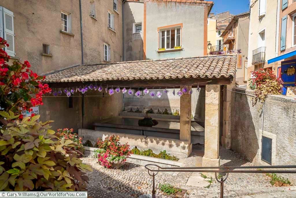 Historic stone lavoir in Valensole, Provence, framed by colorful flowers and pastel village houses under a tiled roof. This traditional Provençal washhouse offers a glimpse into local heritage and adds rustic charm to one of the region’s most picturesque villages.