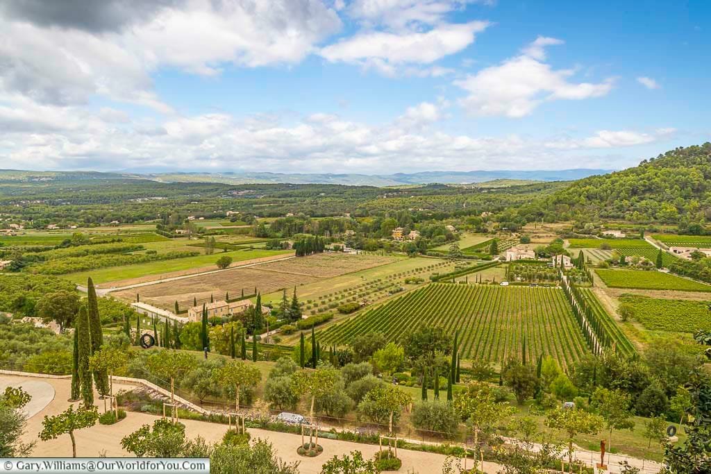 A stunning panoramic view over the Luberon Valley from Ménerbes, showcasing lush vineyards, olive groves, and rolling hills under a partly cloudy Provençal sky. This scenic landscape captures the timeless beauty and agricultural richness of southern France.