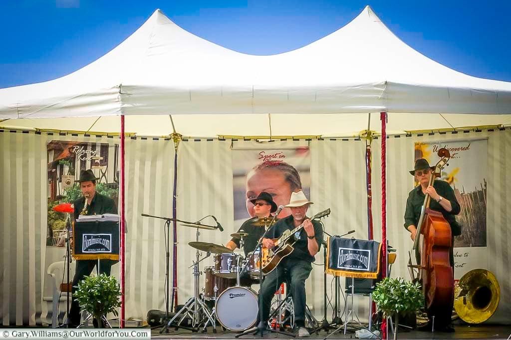 A live jazz band performs under a white canopy at the Kent County Show, bringing a vibrant musical atmosphere to the summer event. The musicians play double bass, drums, saxophone, and guitar,