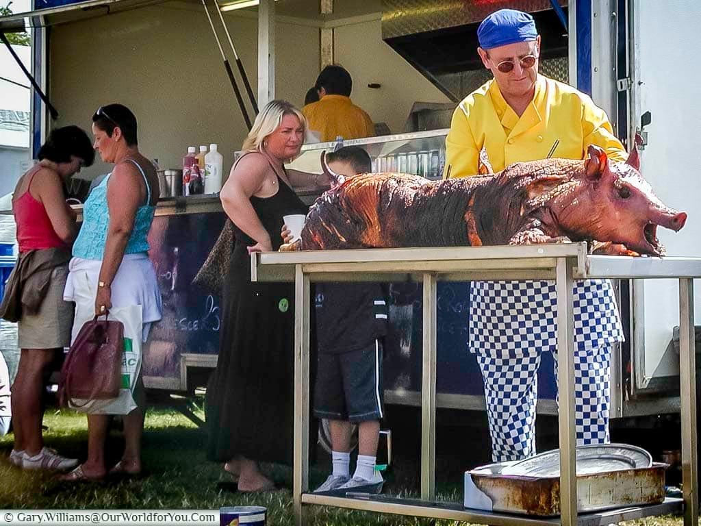 A traditional hog roast being carved by a chef in a bright yellow jacket and checkered trousers at the Kent County Show. Visitors queue at the food stall, enjoying the smells and sights of classic English rural cuisine.