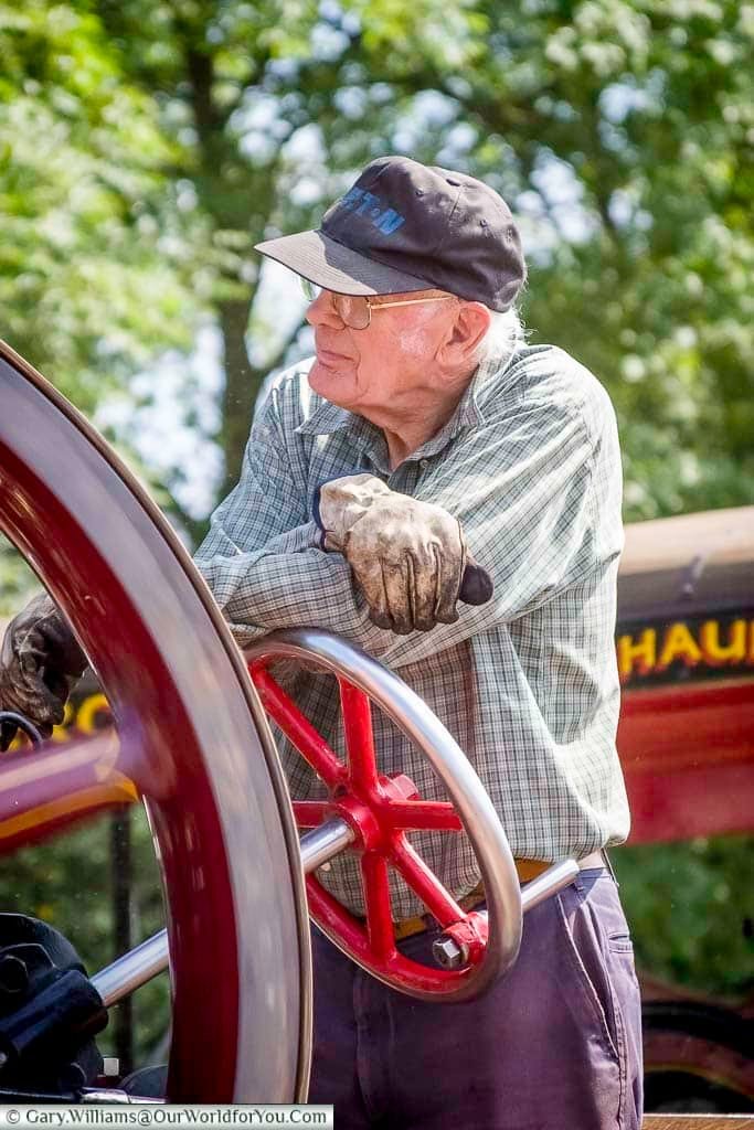 An elderly gentleman leans on a red and silver wheel of a vintage traction engine, taking a break during the Kent County Show. His weathered gloves and relaxed stance evoke the heritage and craftsmanship celebrated at UK rural events.