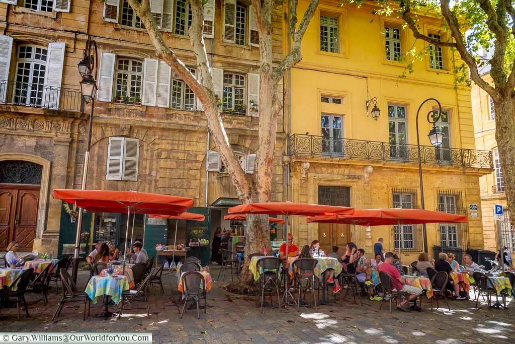 Locals and visitors enjoy outdoor dining at a shady brasserie in Aix-en-Provence, under the wide canopies of orange parasols. The historic façades and relaxed café culture create an authentic Provençal experience.