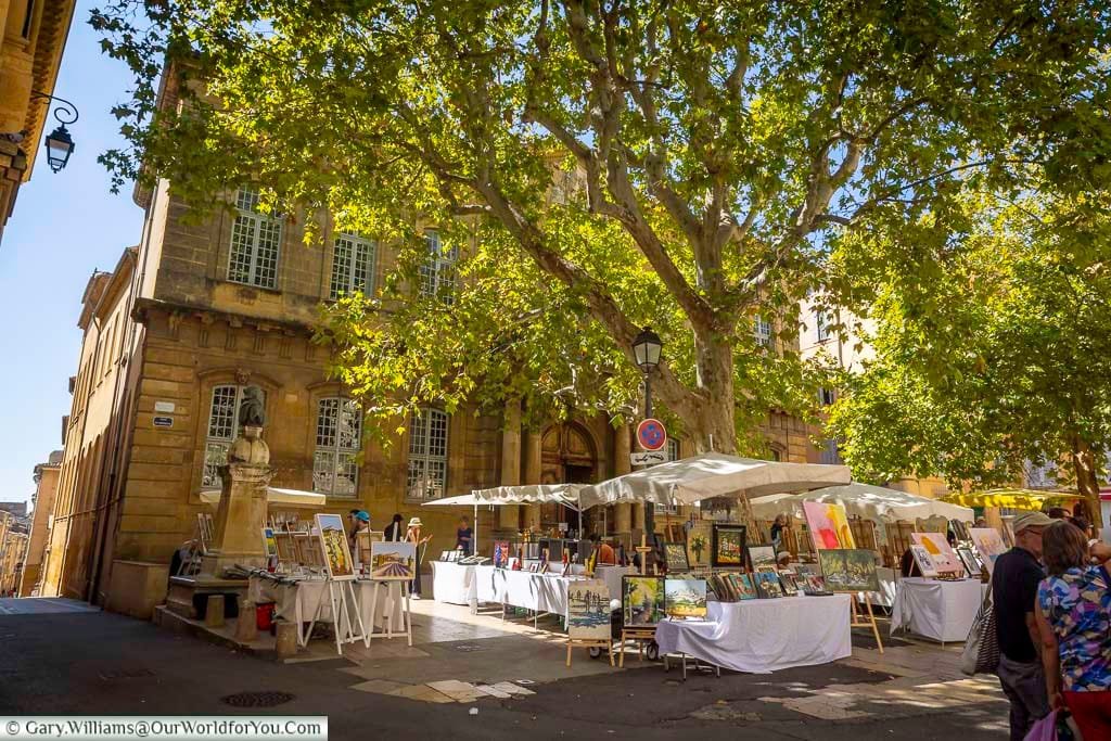 Sunlight filters through plane trees onto a colourful open-air art market in Aix-en-Provence, where local artists exhibit their work. This lively Provençal scene is set against a backdrop of golden stone buildings and fountains.