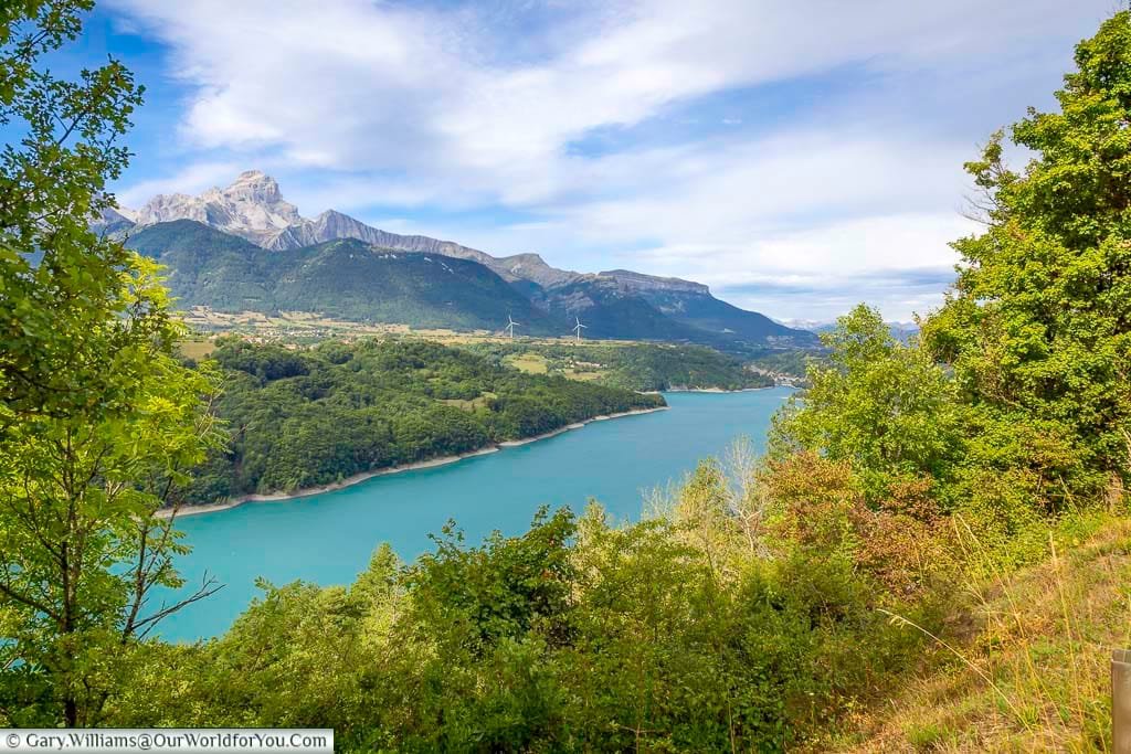 A scenic view of the turquoise waters of Lac de Serre-Ponçon framed by lush greenery, with the dramatic Alpine peaks rising in the distance. This picturesque stop on the way to Sisteron showcases the natural beauty of southeastern France.