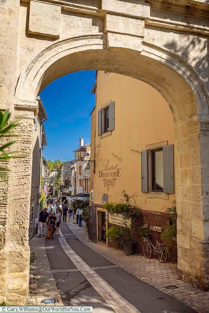 Visitors stroll beneath the historic Porte Saint-Paul archway into the bustling heart of Saint-Rémy-de-Provence. The inviting scene is alive with cafés, wine bars, and Provençal charm in the southern French sunshine.