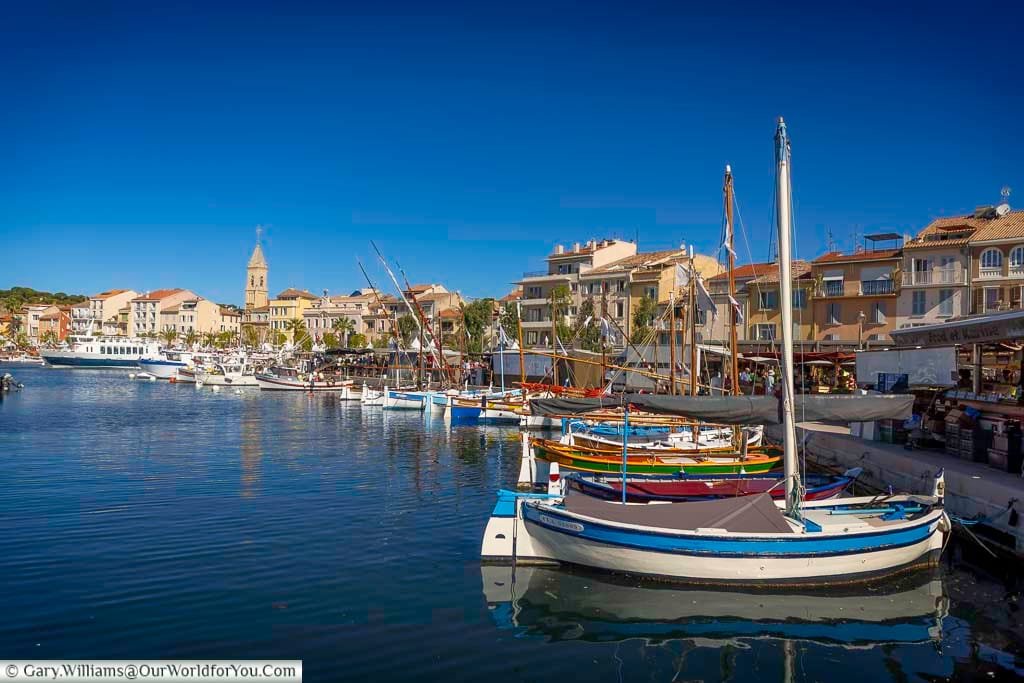 Colourful traditional fishing boats line the glistening bay of Sanary-sur-Mer, with pastel-hued buildings and a church steeple in the background. This lively port on the Mediterranean coast is a stunning stop on any French road trip.