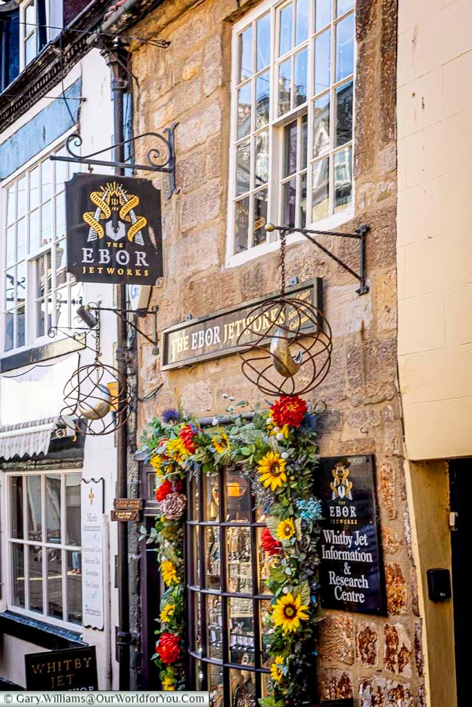 The Ebor Jetworks on Church Street in Whitby features a vibrant flower-framed window and charming hanging sign. Known for its unique jewellery made from Whitby jet, this shop is a must-visit along the town's historic lanes.