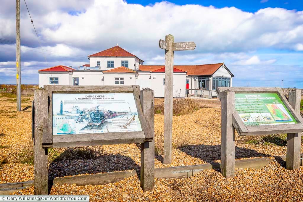 A view of the visitor information boards at the Dungeness National Nature Reserve, with a signpost directing visitors to the sea and a white building with a red roof in the background under a dramatic sky.