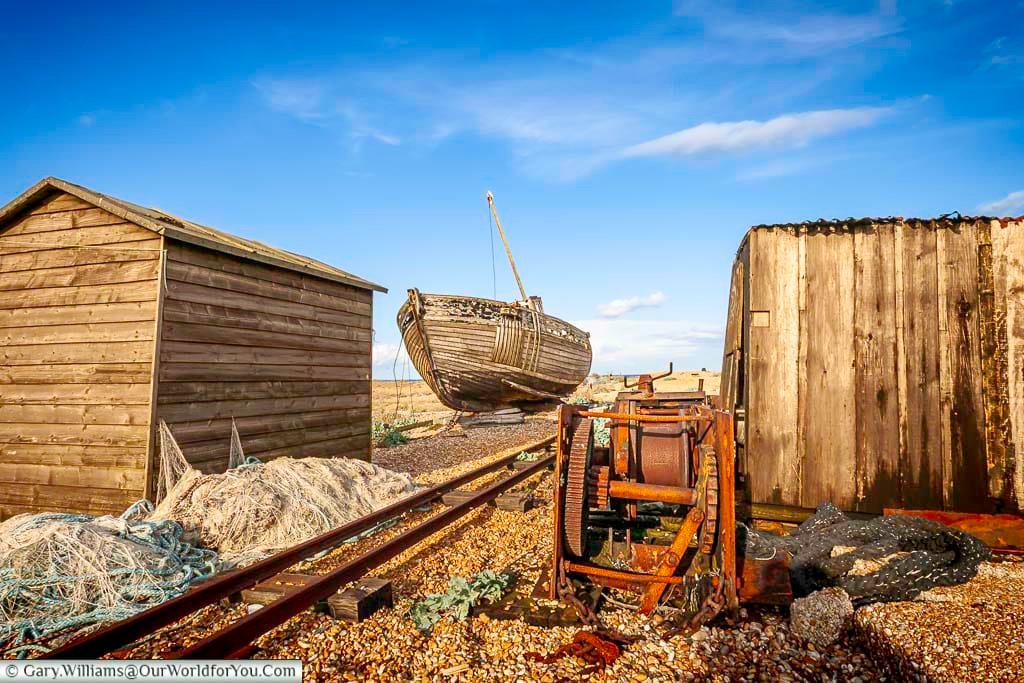 An atmospheric scene of weather-beaten sheds, a rusted winch, and an abandoned fishing boat resting on rail tracks, echoing Dungeness’s industrial fishing heritage. This evocative image captures the raw, unpolished charm of this remote Kent headland.