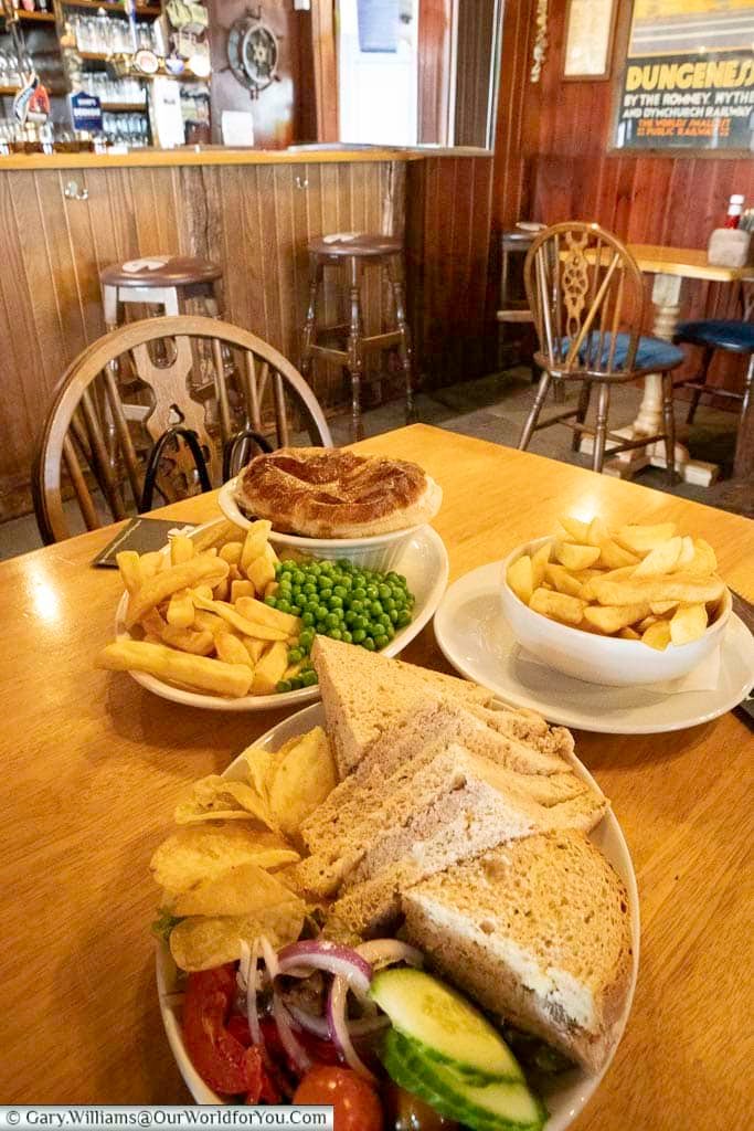 A hearty British pub lunch at the Pilot Inn in Dungeness, featuring a traditional steak pie with chips and peas, alongside a sandwich platter with crisps and salad. Set in a rustic coastal pub, this classic Kentish meal offers a warm welcome after exploring the windswept Dungeness headland.