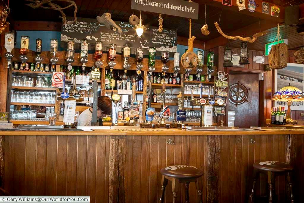 The warm and inviting bar of the Pilot Inn in Dungeness, lined with spirits, beer taps, and a charming mix of driftwood and seafaring memorabilia. A traditional Kentish pub offering local hospitality by the coast.