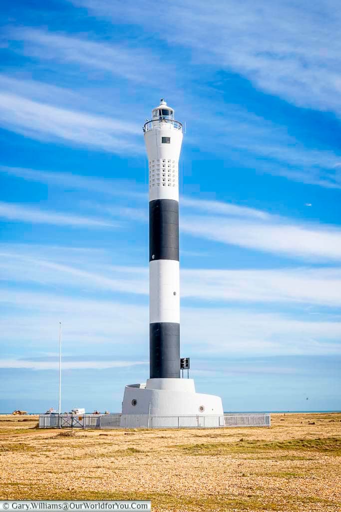 The modern black-and-white striped Dungeness lighthouse stands tall under blue Kentish skies, continuing the legacy of coastal navigation along this rugged shoreline. It contrasts sharply with the surrounding flat landscape of shingle and sky.