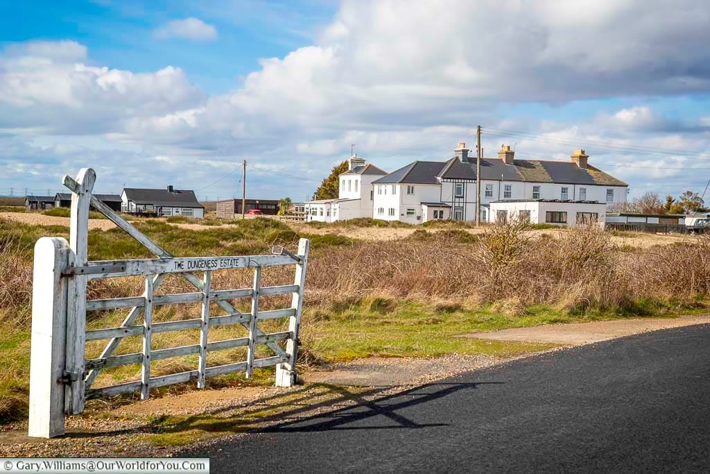 A weathered white gate marks the boundary of the remote Dungeness Estate in Kent, where stark beauty and wild landscapes meet the sea. Behind it, historic cottages line the horizon, shaped by wind and time.