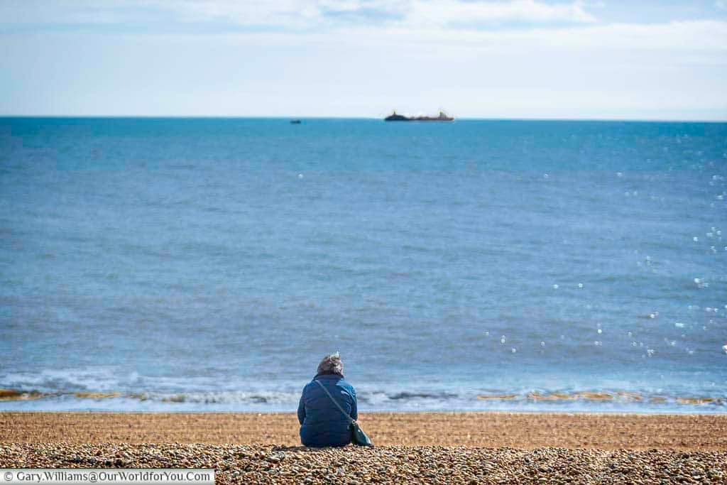 A lone figure sits on the shingle beach at Dungeness, gazing out to the distant ships across the calm, blue English Channel. This peaceful scene evokes quiet contemplation amidst the expansive coastal landscape.