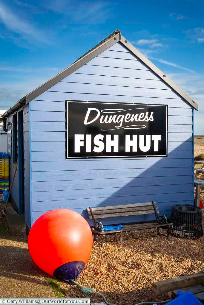 A bright blue wooden hut with a bold “Dungeness Fish Hut” sign stands on the shingle beach under a clear Kent sky. This iconic spot on the Dungeness coast is where visitors can buy freshly caught seafood straight from the local fishing fleet.