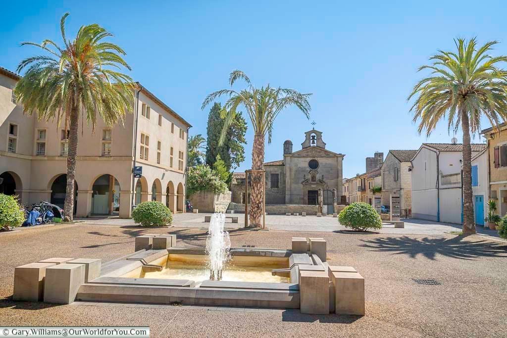 Palm trees and fountains frame Place de la Viguerie in Aigues-Mortes, with the 17th-century Église Notre-Dame-des-Sablons anchoring the square. This peaceful plaza is a serene spot in the heart of this historic Occitanie town.