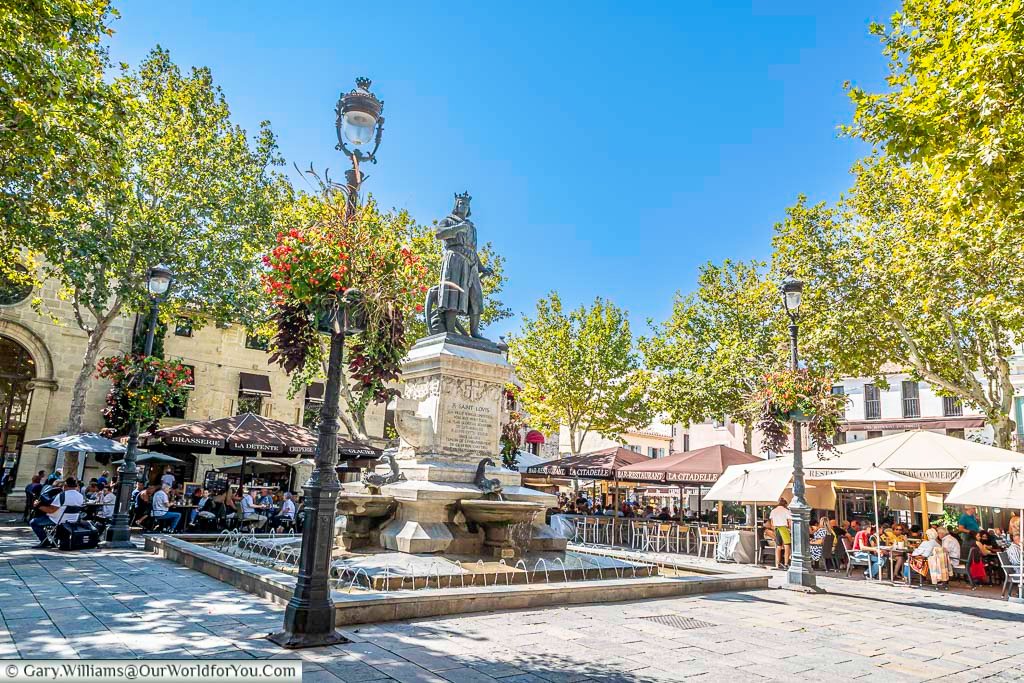 A lively summer scene at Place Saint Louis in Aigues-Mortes, filled with café terraces, hanging baskets, and a central fountain beneath blue skies. This historic square in Occitanie is a must-see destination for travellers exploring southern France.
