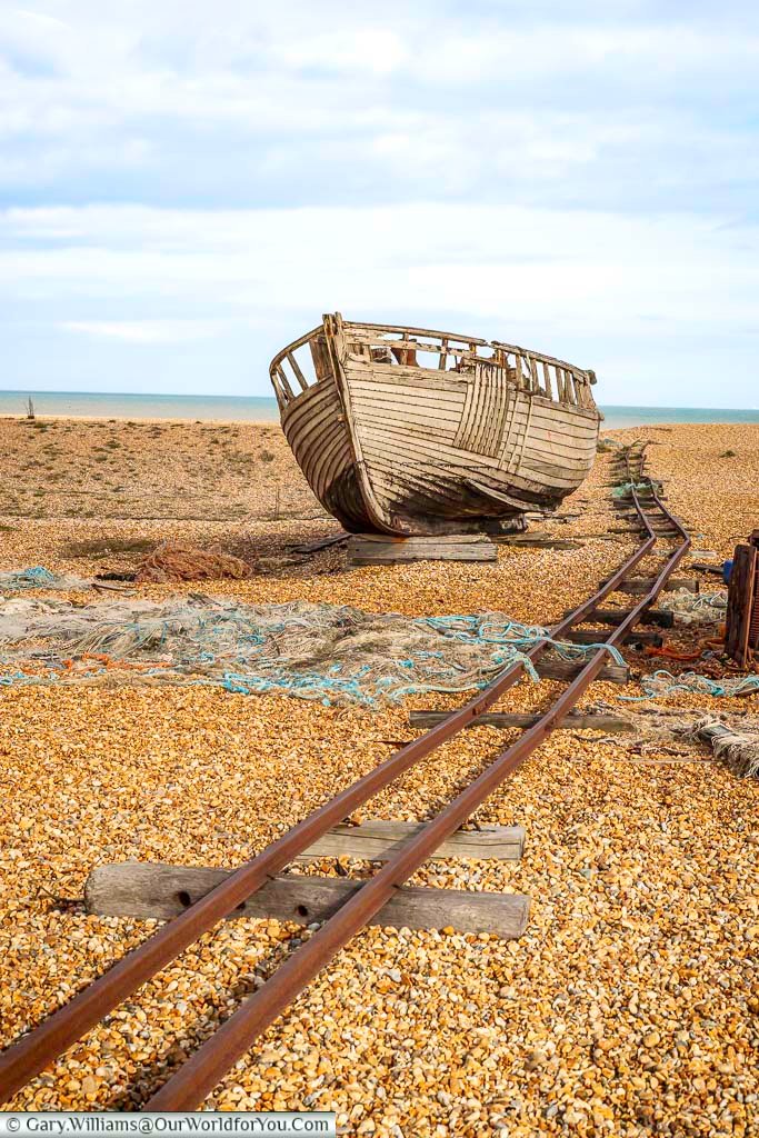 A weathered wooden fishing boat rests beside rusted rail tracks on the shingle beach at Dungeness, Kent, under a bright coastal sky. This evocative scene captures the raw beauty and heritage of England’s most unusual landscape, a must-visit for photographers exploring hidden gems along the Kent coast.