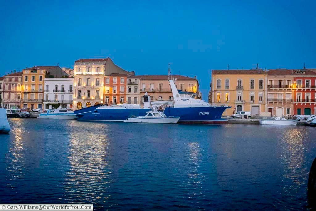 As the evening light fades, the pastel-coloured buildings of Sète glow warmly across the harbourfront. Luxury yachts and fishing boats gently bob in the tranquil blue waters, capturing the essence of a Mediterranean dusk.