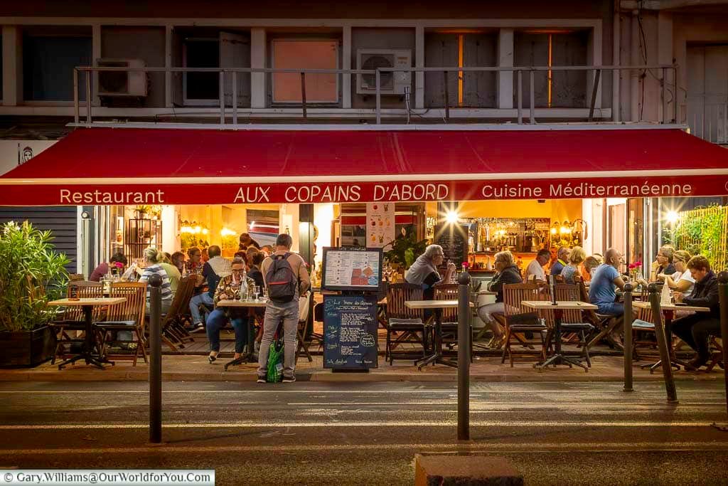 Diners gather at the lively Aux Copains d’Abord restaurant in Sète, enjoying Mediterranean cuisine under a glowing red awning. The evening ambiance and friendly buzz highlight the city’s vibrant nightlife and coastal charm.