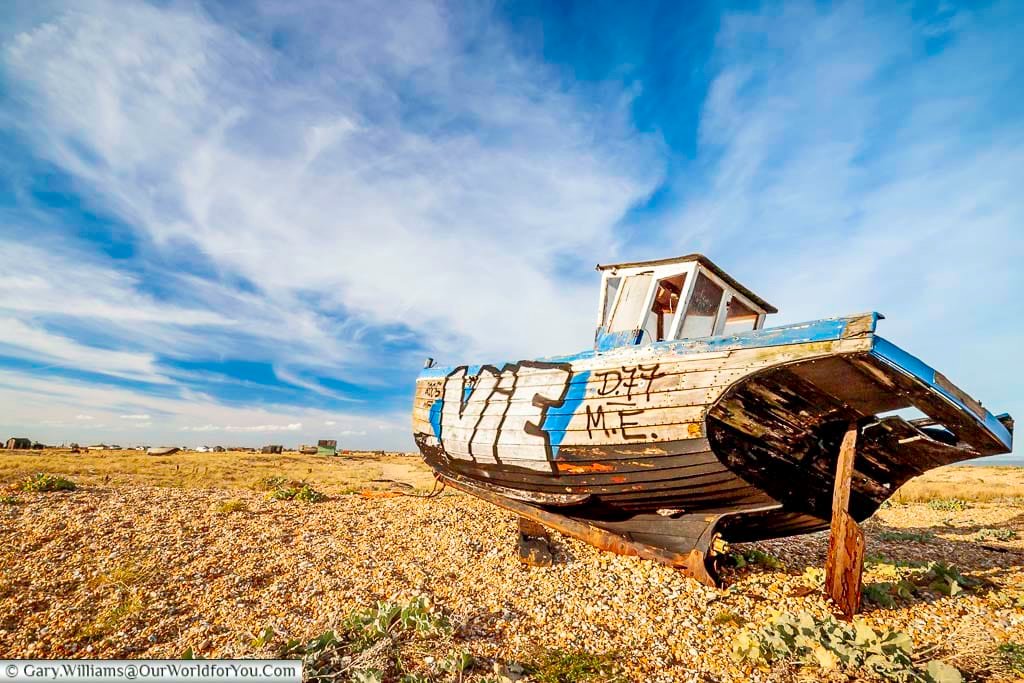 A weathered fishing boat with blue paint and graffiti sits abandoned on the shingle beach at Dungeness, Kent, beneath a wide, cloud-streaked sky. This early image captures the raw, artistic character of Dungeness before time and tide began to erode its iconic relics.