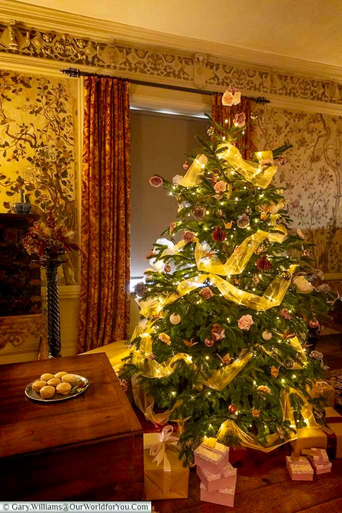 A beautifully decorated Christmas tree stands in the Drawing Room at Ightham Mote, surrounded by historic furnishings and soft golden lighting. The scene captures the festive spirit of this medieval Kent manor.