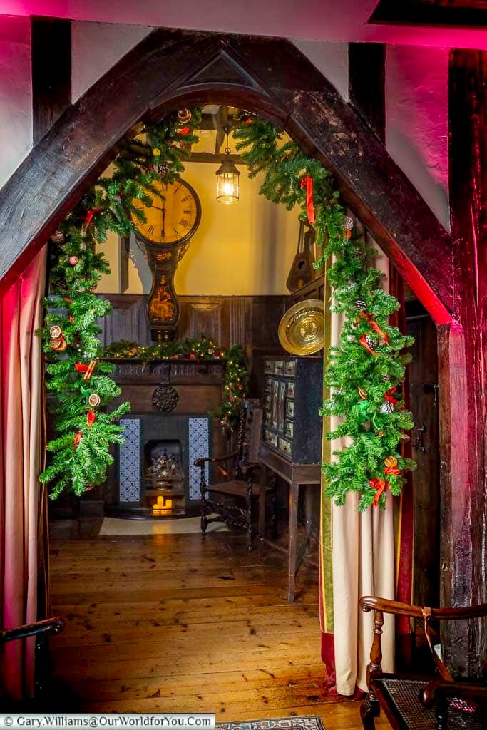 A garland-framed doorway leads through a candlelit corridor inside Ightham Mote, decorated for Christmas with seasonal greenery and soft lighting. The medieval passageway highlights the historic atmosphere of this much-loved Kent landmark.