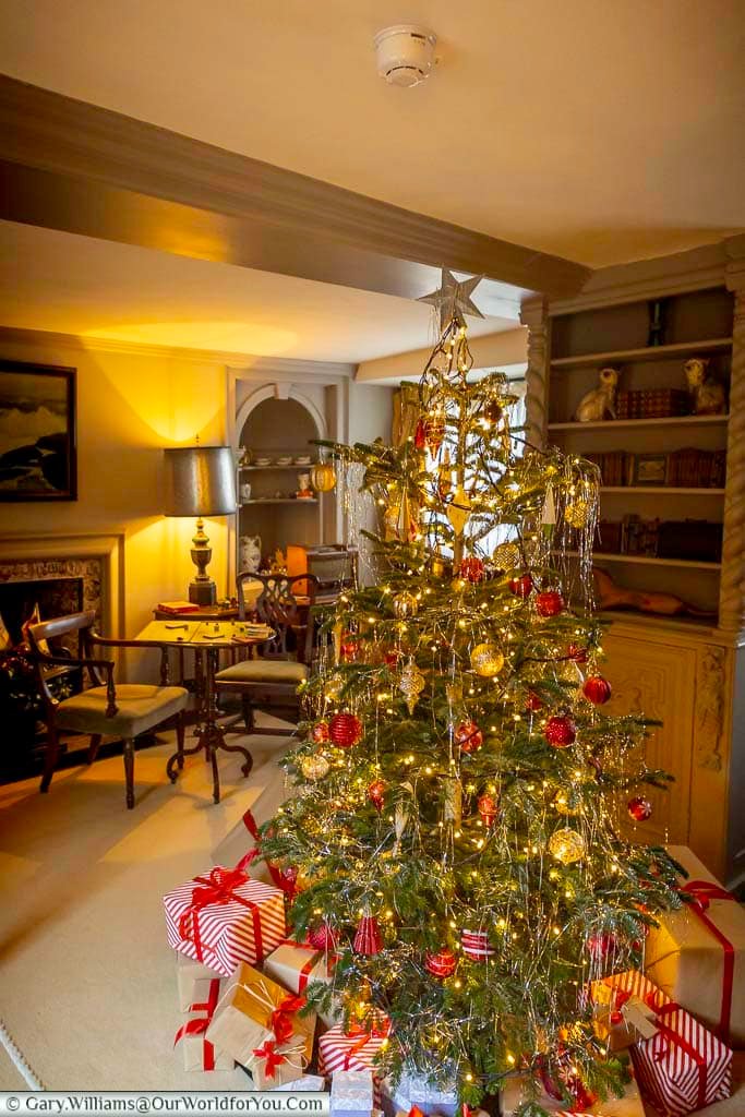 A fully decorated Christmas tree stands among bookshelves and period furniture in the library at Ightham Mote. Wrapped presents and warm lighting create a nostalgic festive atmosphere.