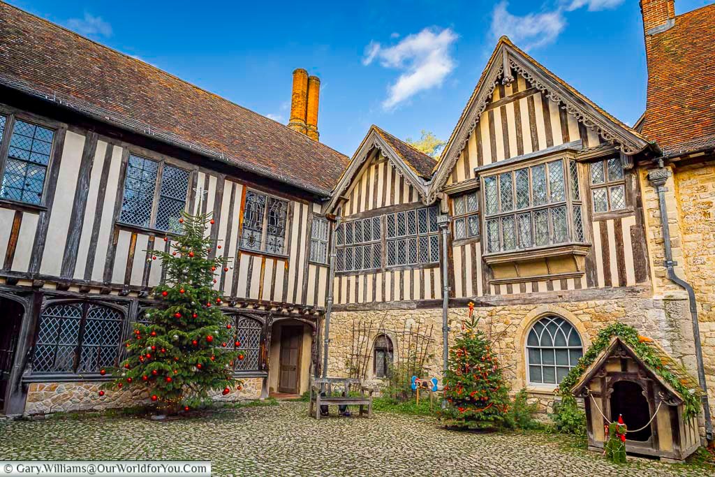 A festive view across the courtyard at Ightham Mote, showing decorated Christmas trees and the charming historic dog kennel beside the stone walls. This seasonal scene captures the character of this medieval Kent manor and its unique traditions within the National Trust estate.