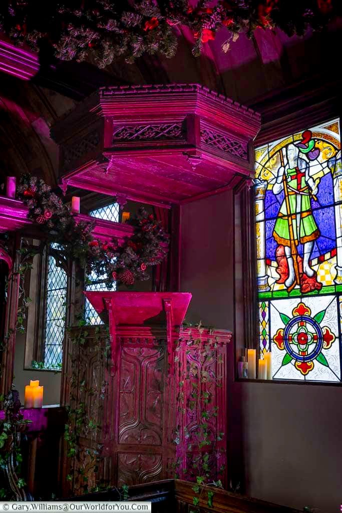The ornately carved wooden pulpit inside the New Chapel at Ightham Mote in Kent, illuminated with atmospheric lighting and framed by a colourful stained-glass window. This striking detail highlights the craftsmanship and medieval heritage of this National Trust property, making it a must-see feature when exploring historic places in Kent.
