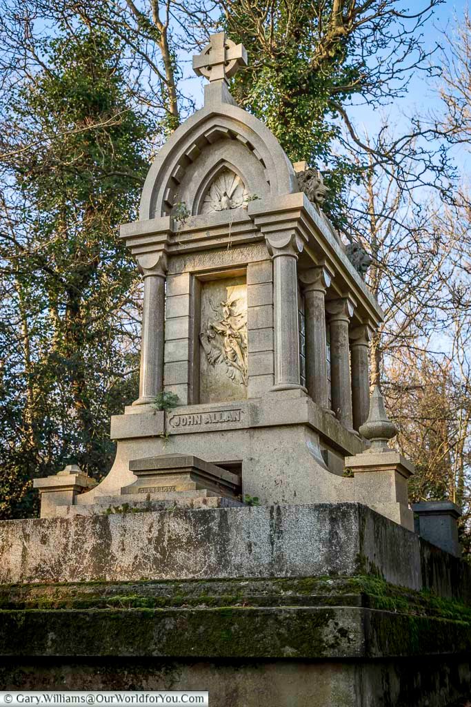 Ornate stone tomb of John Allan featuring carved religious reliefs and Gothic detailing in Nunhead Cemetery, London. This historic Victorian monument is surrounded by mature trees, highlighting the cemetery’s architectural heritage and tranquil setting.