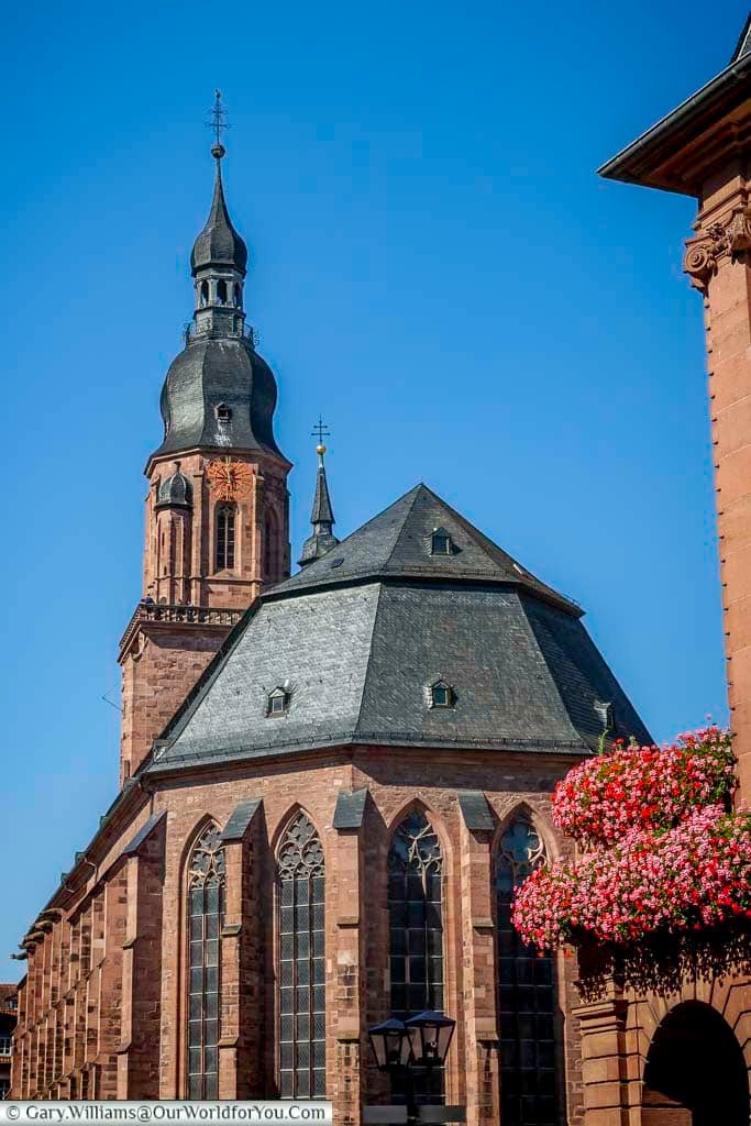 Side view of the gothic nave and tower of the Church of the Holy Spirit with flower-filled balconies in the foreground. The red sandstone walls and tall arched windows emphasise the rich architectural heritage of historic Heidelberg.
