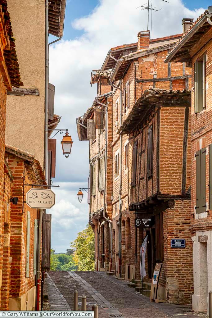 Traditional brick street in Albi with hanging signs, shuttered windows, and a glimpse toward the River Tarn. This classic streetscape perfectly captures the historic charm of Albi, making it a highlight of any French road trip.