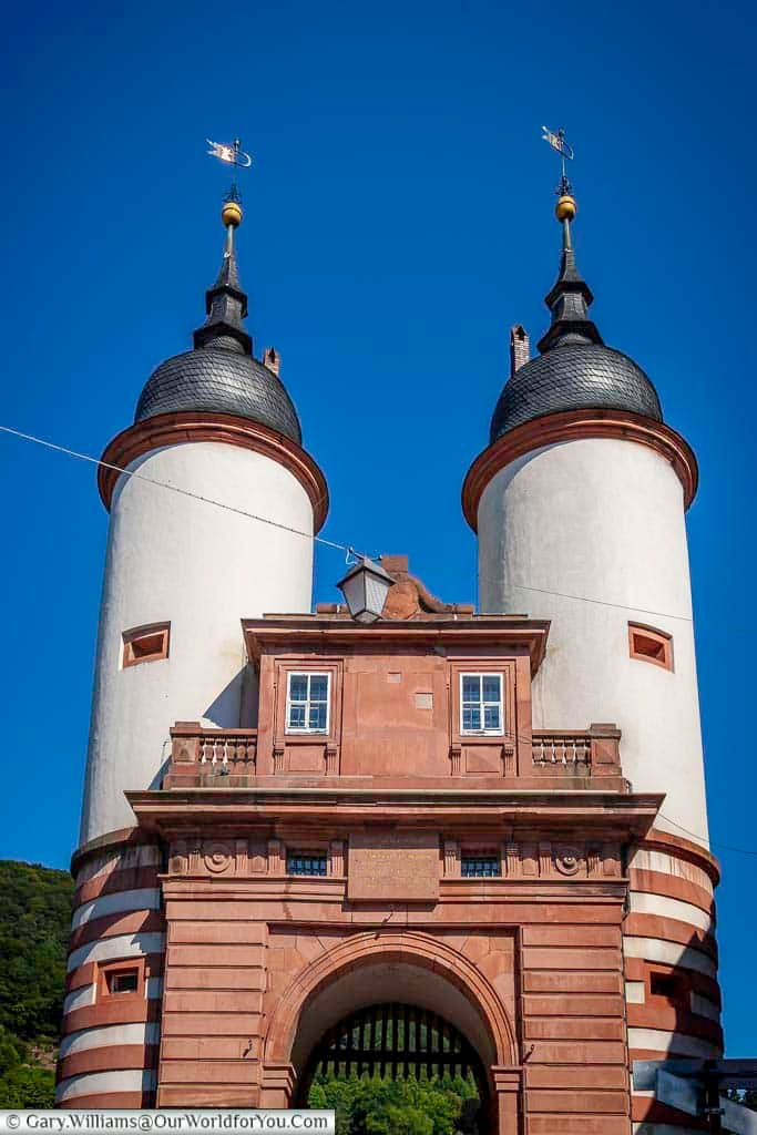 Twin white towers of the Old Bridge Gate marking the entrance to Heidelberg’s historic Alte Brücke under a clear blue sky. Iconic red sandstone gateway connecting Heidelberg Old Town with panoramic views across the River Neckar.