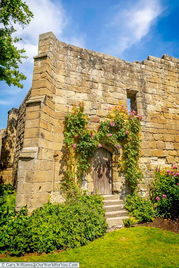 A charming stone doorway covered in blooming pink roses at Mount Grace Priory, a National Trust site in North Yorkshire. This historic entrance is surrounded by lush greenery and medieval stonework under a bright summer sky.