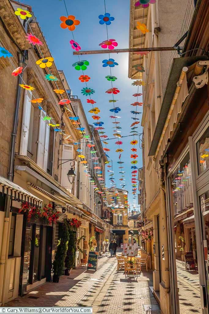 Bright hanging flower decorations fill the sky above a narrow shopping street in Bergerac, creating a cheerful and picturesque scene. This charming view showcases the lively atmosphere of the town centre in Dordogne, France.