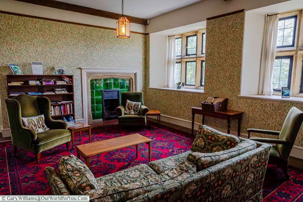 A cosy historic sitting room inside Mount Grace House, featuring vintage furniture, patterned wallpaper, and a striking green tiled fireplace. This beautifully restored National Trust property in North Yorkshire showcases traditional English country house style.