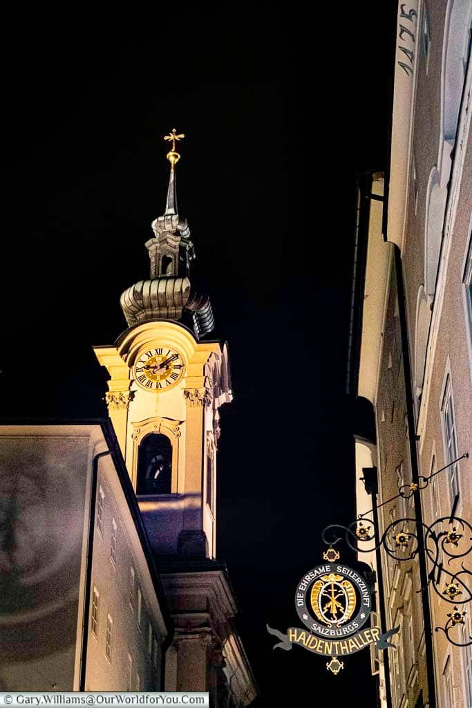 Linzer Gasse in Salzburg photographed at night, with illuminated shop signs and church towers glowing against the dark sky. This atmospheric street is known for its historic charm, local shops, and evening strolls.