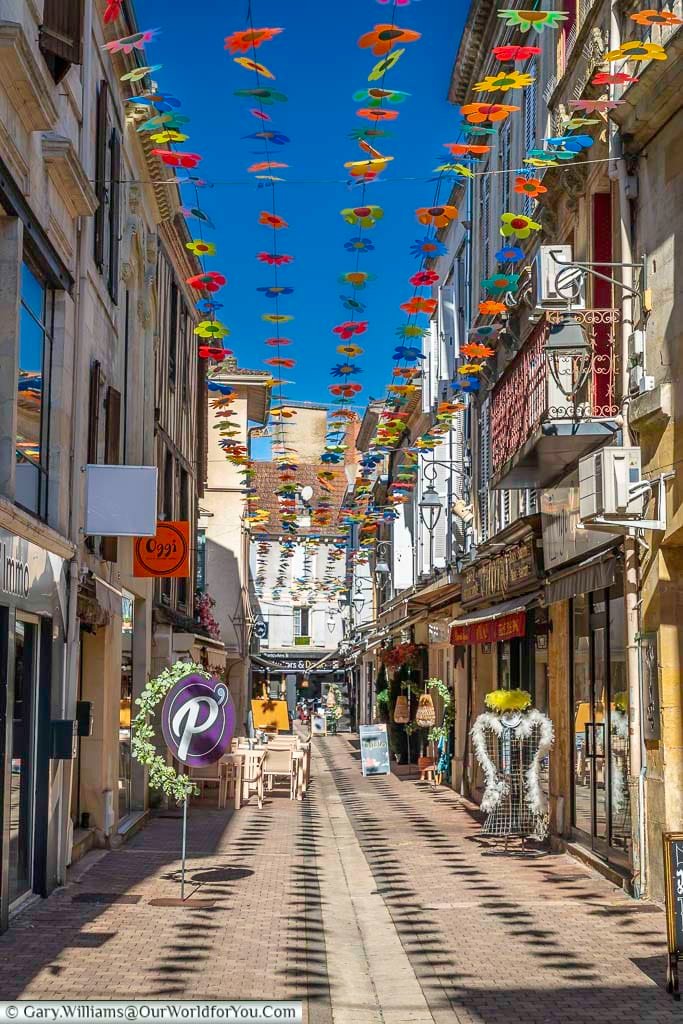 Colourful flower decorations hang above Rue du Mourrier in Bergerac, creating a vibrant canopy over the charming pedestrian street. This lively scene captures the atmosphere of the historic town centre in the Dordogne region of France.