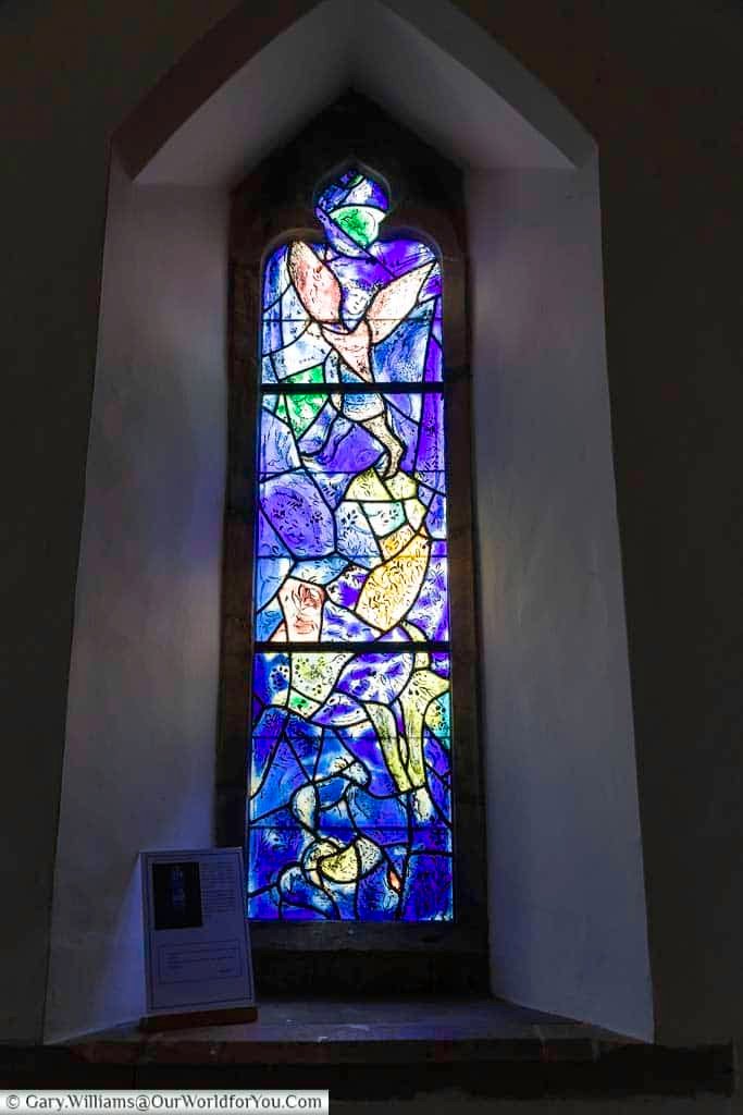 A colourful stained glass window by Marc Chagall features an abstract angel and symbolic figures, including personal motifs, in All Saints’ Church, Tudeley. The intricate design reflects the emotional story behind this unique Kent landmark.
