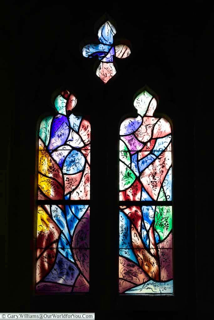 A warm-toned stained glass window by Marc Chagall features abstract figures and a distinctive blue fish within a quatrefoil design at Tudeley Church. The glowing colours create a beautiful contrast within this historic Kent interior.