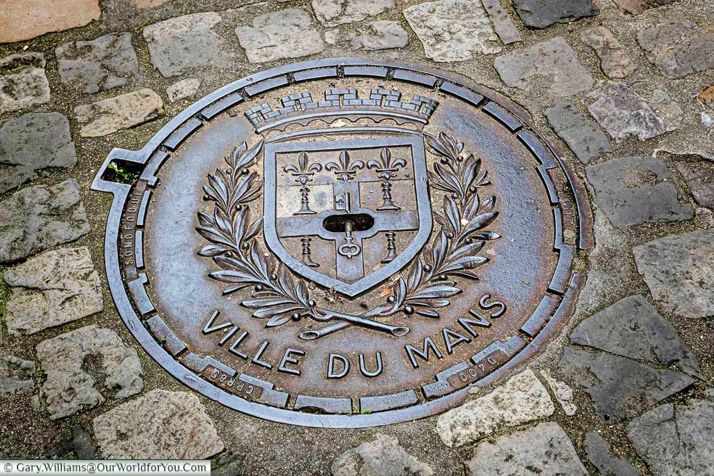 A detailed “Ville du Mans” coat of arms is embedded in a cobbled street, surrounded by historic paving stones. This decorative manhole cover reflects the city’s heritage and adds charm to the streets of Le Mans.