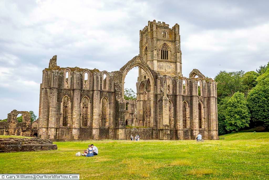 Expansive exterior view of Fountains Abbey ruins in Yorkshire, with visitors relaxing on the grass beside the towering medieval structure. The centuries-old abbey is one of the most famous historic sites in England and a highlight of exploring hidden Yorkshire landmarks.