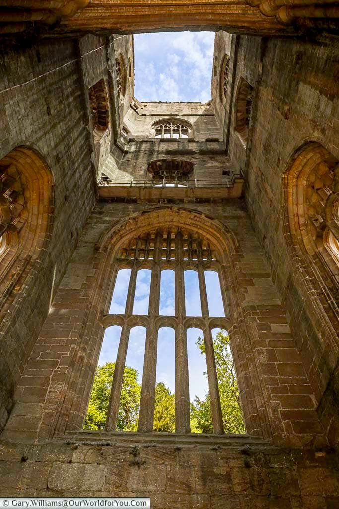 Looking up inside Huby’s Tower at Fountains Abbey, with towering Gothic stone walls and tall medieval windows framing the blue sky above. The dramatic architecture makes this hidden Yorkshire landmark one of the most fascinating historic places to explore in England.