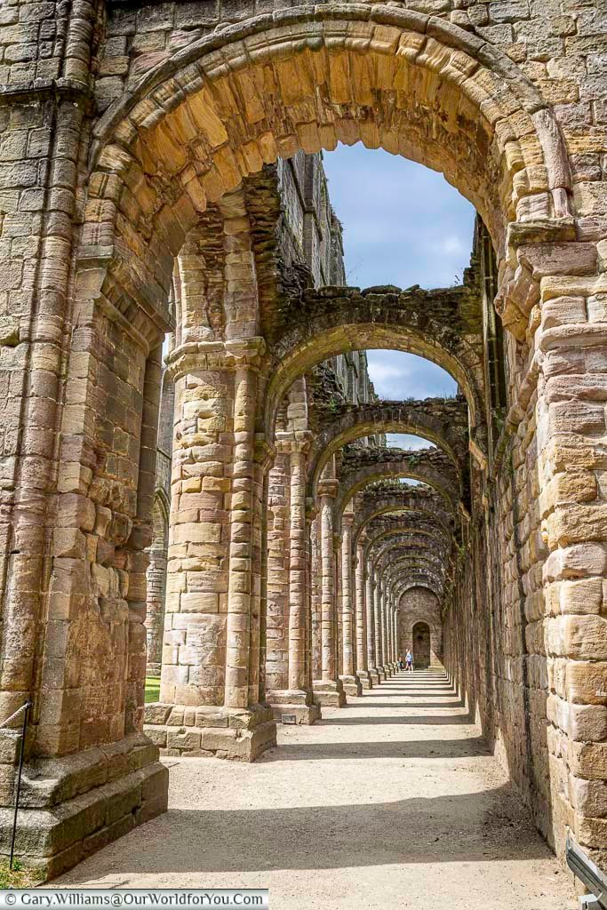 Sunlight filtering through the long stone arches inside the nave of Fountains Abbey, creating dramatic shadows along the ancient medieval corridor. This historic Yorkshire landmark offers visitors a breathtaking glimpse into the grandeur of one of England’s best-preserved abbey ruins.