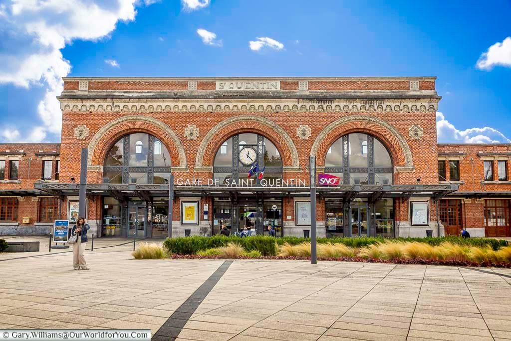 The historic Saint-Quentin train station features a striking red-brick façade with elegant arches and classic French railway architecture beneath bright summer skies. This beautifully restored SNCF station is an important gateway for visitors exploring Saint-Quentin, Hauts-de-France, and scenic French rail journeys.