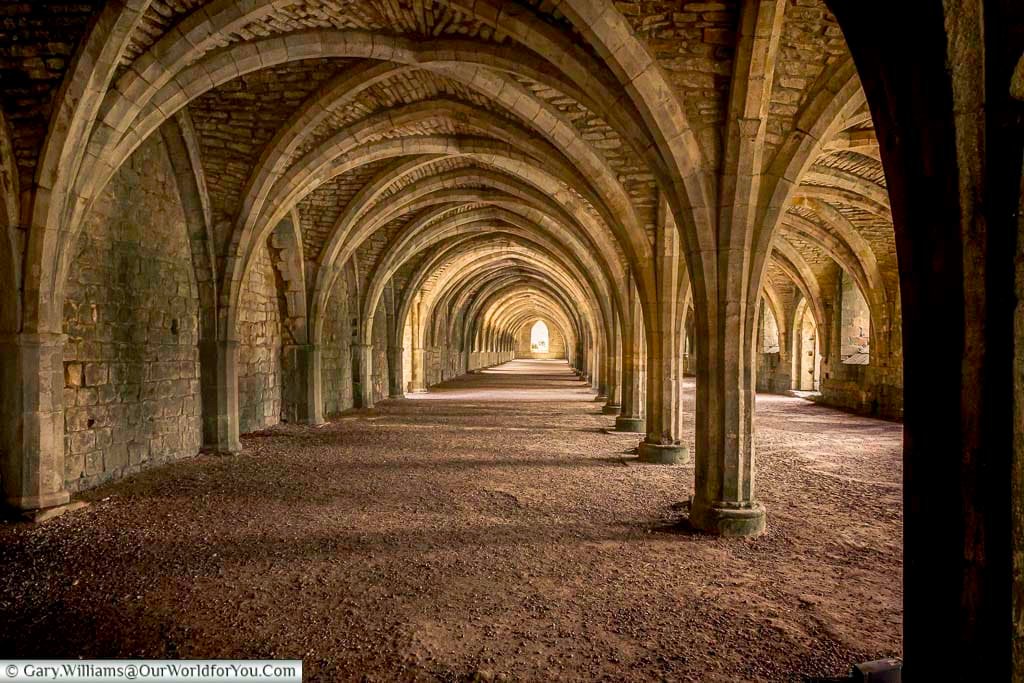 Long vaulted cellarium at Fountains Abbey in Yorkshire, featuring repeating medieval stone arches and dramatic light streaming through the ancient corridor. This impressive monastic storage hall highlights the scale and craftsmanship of one of England’s best-preserved abbey ruins.